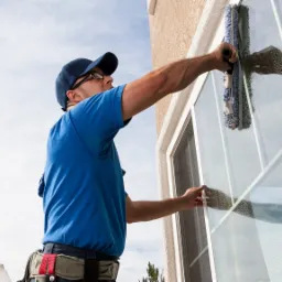 Pracownik w niebieskiej koszulce i czapce, z pasem narzędzi, myje okno szklaną ściągaczką, widoczne smugi wody na szybie i odbicie sylwetki pracownika.
