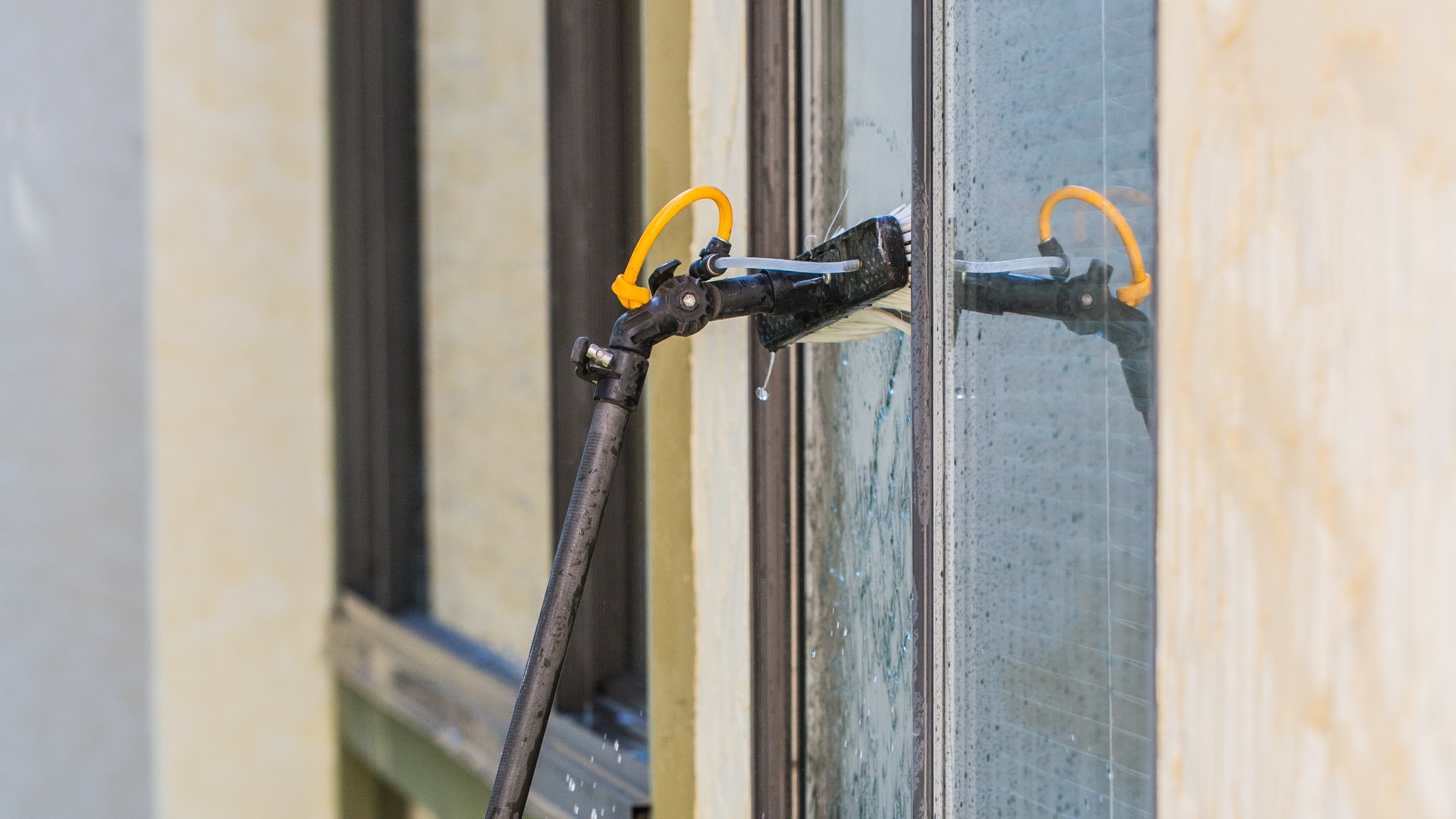Szczotka na teleskopowym kiju z żółtym przewodem myje okno, woda spływa po szybie, widoczna rama okienna i fragment elewacji budynku.