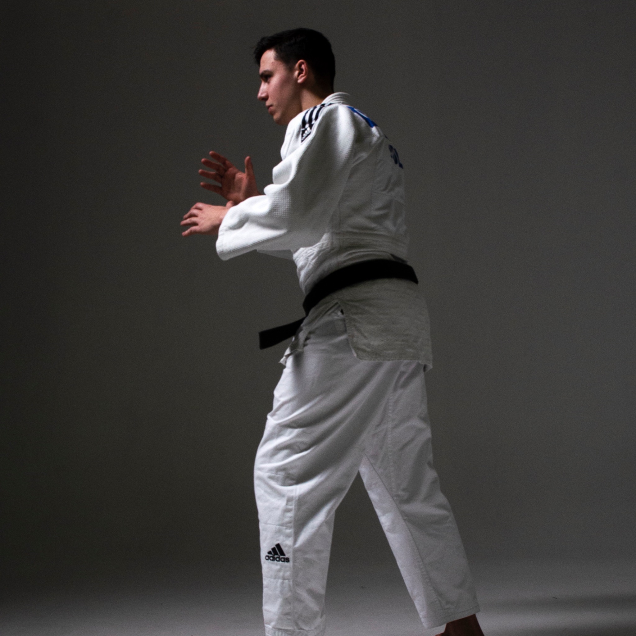 Młody mężczyzna w białym judogi z czarnym pasem, w pozycji gotowości do walki, profil, logo Adidas na spodniach.