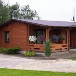 Niski, parterowy budynek o drewnianej konstrukcji z zadaszonym tarasem, ozdobiony wiszącymi kwiatami, otoczony zielenią i niskimi iglakami. Do budynku prowadzi betonowa droga.