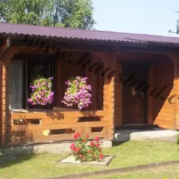 Parterowy budynek z drewnianych bali z zadaszonym tarasem, ozdobiony wiszącymi koszami z kwiatami i zadbanym trawnikiem z ozdobnymi krzewami.