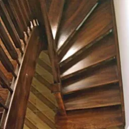 Kręte schody drewniane z ciemnego drewna, z widocznymi poręczami i stopniami, ułożone nad podłogą z wzorem w paski.