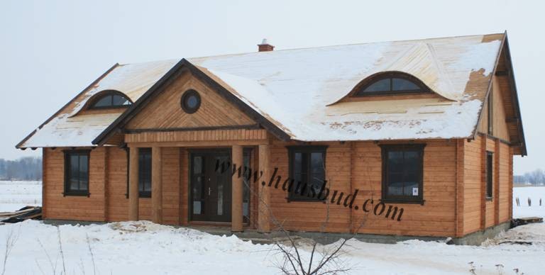 Parterowy dom z jasnego drewna z dachem pokrytym śniegiem, okrągłe i półokrągłe okna na poddaszu, widoczne ślady budowy na śniegu.