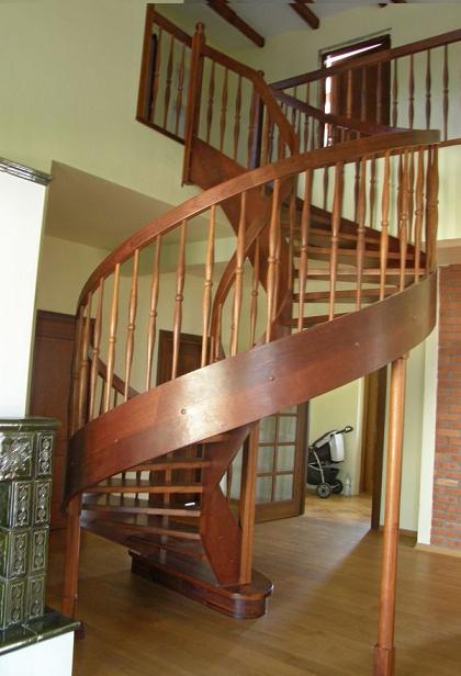Drewniane, spiralne schody z balustradą w domu w Choczu, widoczne z dołu, z fragmentem ozdobnego pieca kaflowego po lewej stronie i wózkiem dziecięcym w tle.
