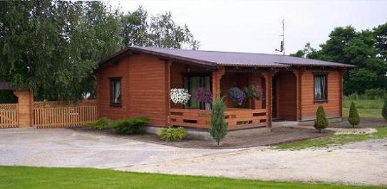 Niski, parterowy budynek o drewnianej konstrukcji z zadaszonym tarasem, ozdobiony wiszącymi kwiatami, otoczony zielenią i niskimi iglakami. Do budynku prowadzi betonowa droga.