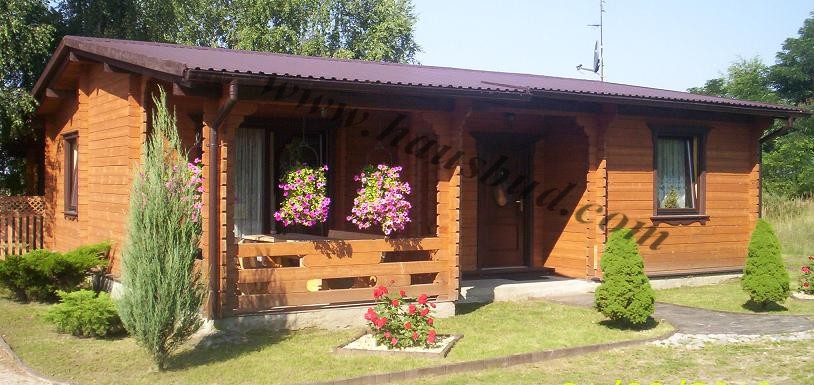 Parterowy budynek z drewnianych bali z zadaszonym tarasem, ozdobiony wiszącymi koszami z kwiatami i zadbanym trawnikiem z ozdobnymi krzewami.