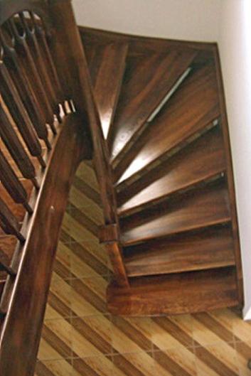 Kręte schody drewniane z ciemnego drewna, z widocznymi poręczami i stopniami, ułożone nad podłogą z wzorem w paski.