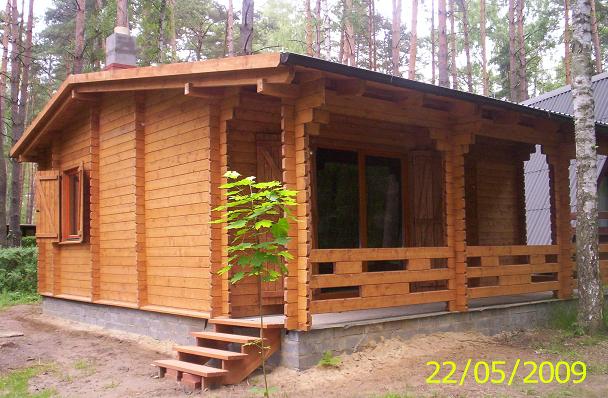 Niewielki, parterowy budynek z jasnego drewna, zadaszony, z drewnianymi okiennicami i schodami wejściowymi, otoczony drzewami.