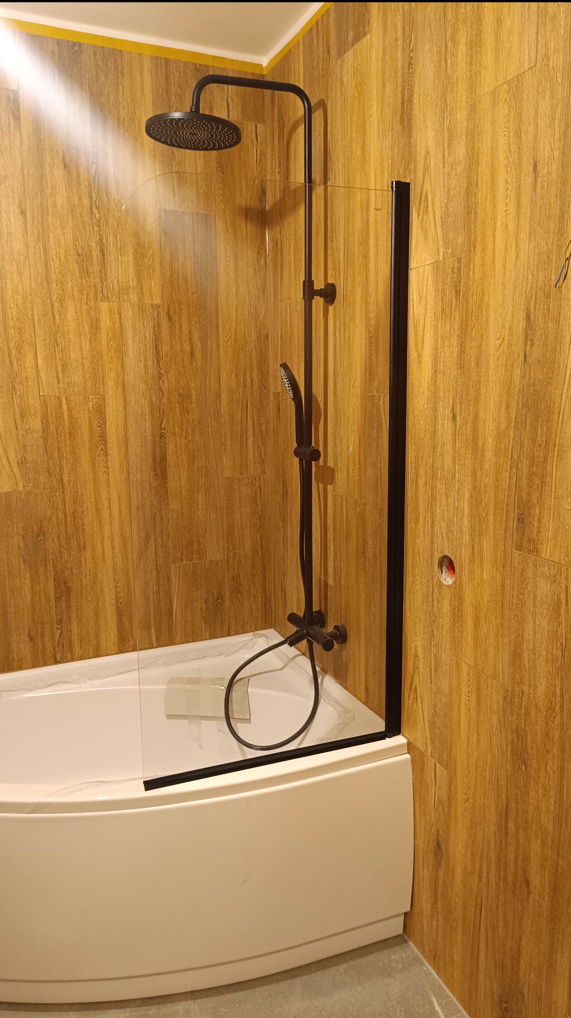 Nowoczesna, czarna armatura prysznicowa zamontowana nad białą wanną narożną, zabezpieczoną folią ochronną, na tle ściany wyłożonej panelami imitującymi drewno.