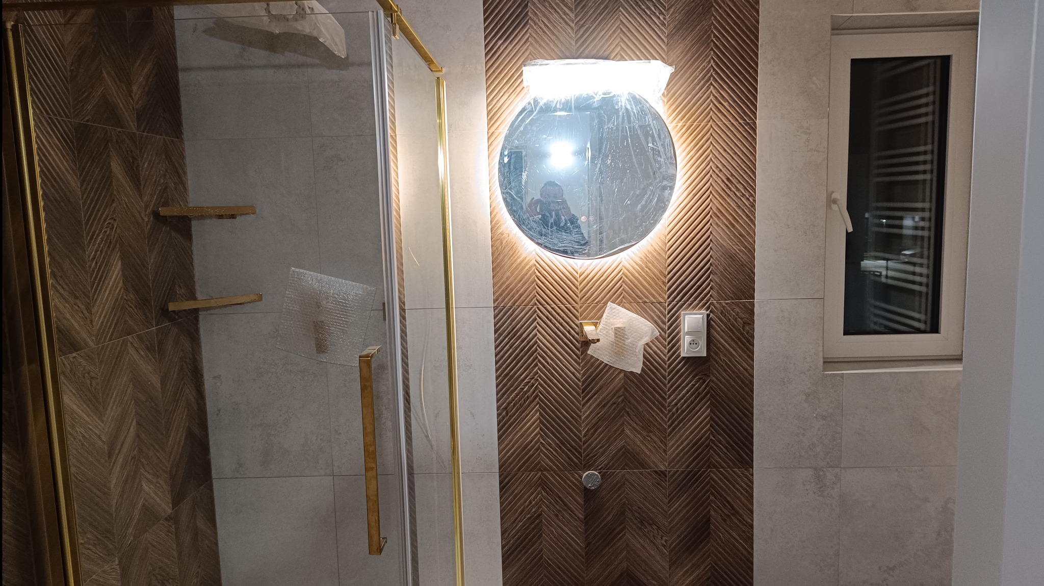 Nowoczesna łazienka w trakcie wykańczania: kabina prysznicowa ze złotymi detalami, okrągłe lustro z podświetleniem LED w folii ochronnej, dekoracyjne płytki imitujące drewno ułożone w jodełkę...