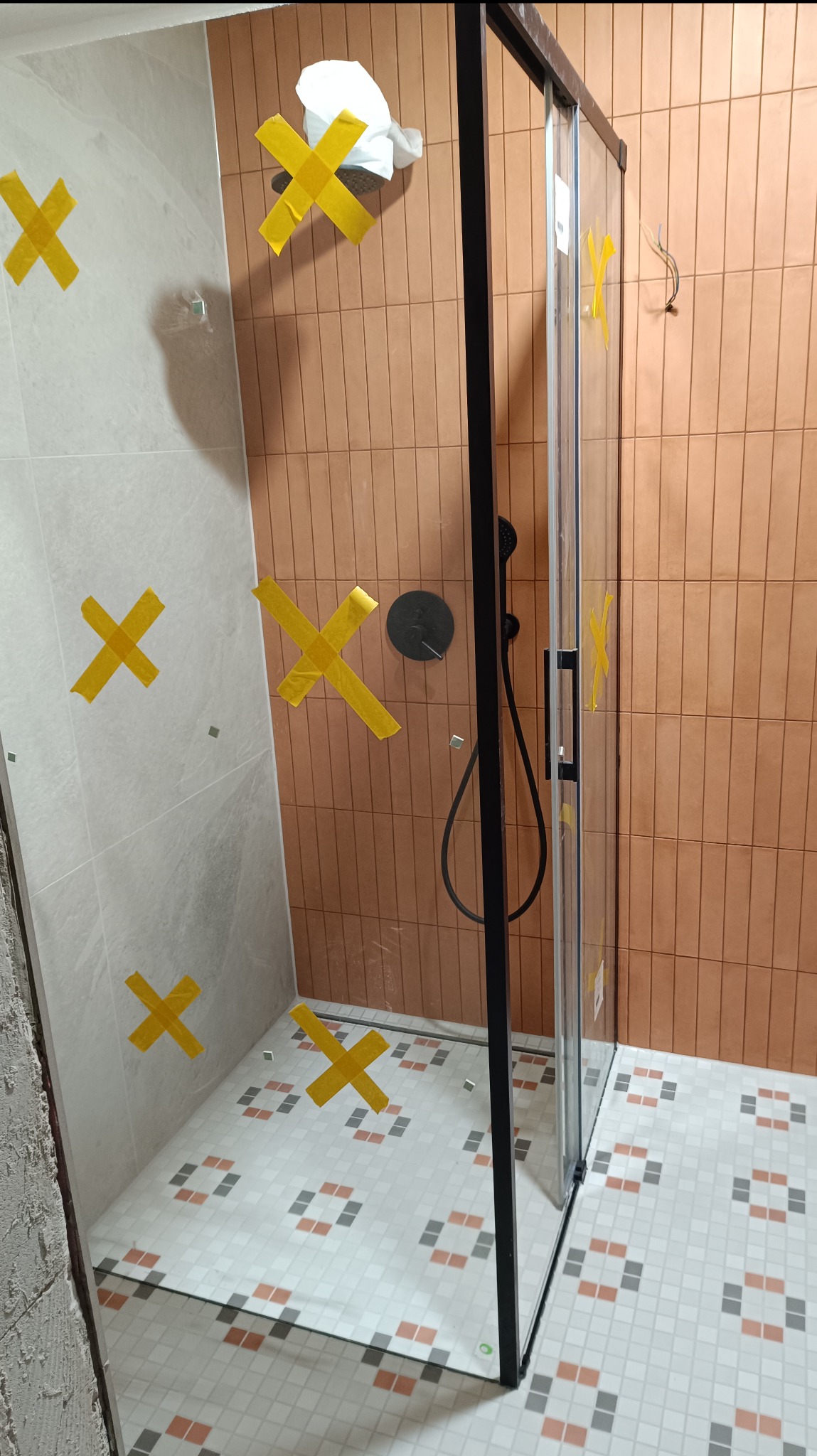 Nowoczesna kabina prysznicowa z czarnymi profilami, zamontowana w łazience z mozaikową podłogą i różnymi rodzajami płytek ściennych, zabezpieczona taśmą malarską na czas prac wykończeniowych.