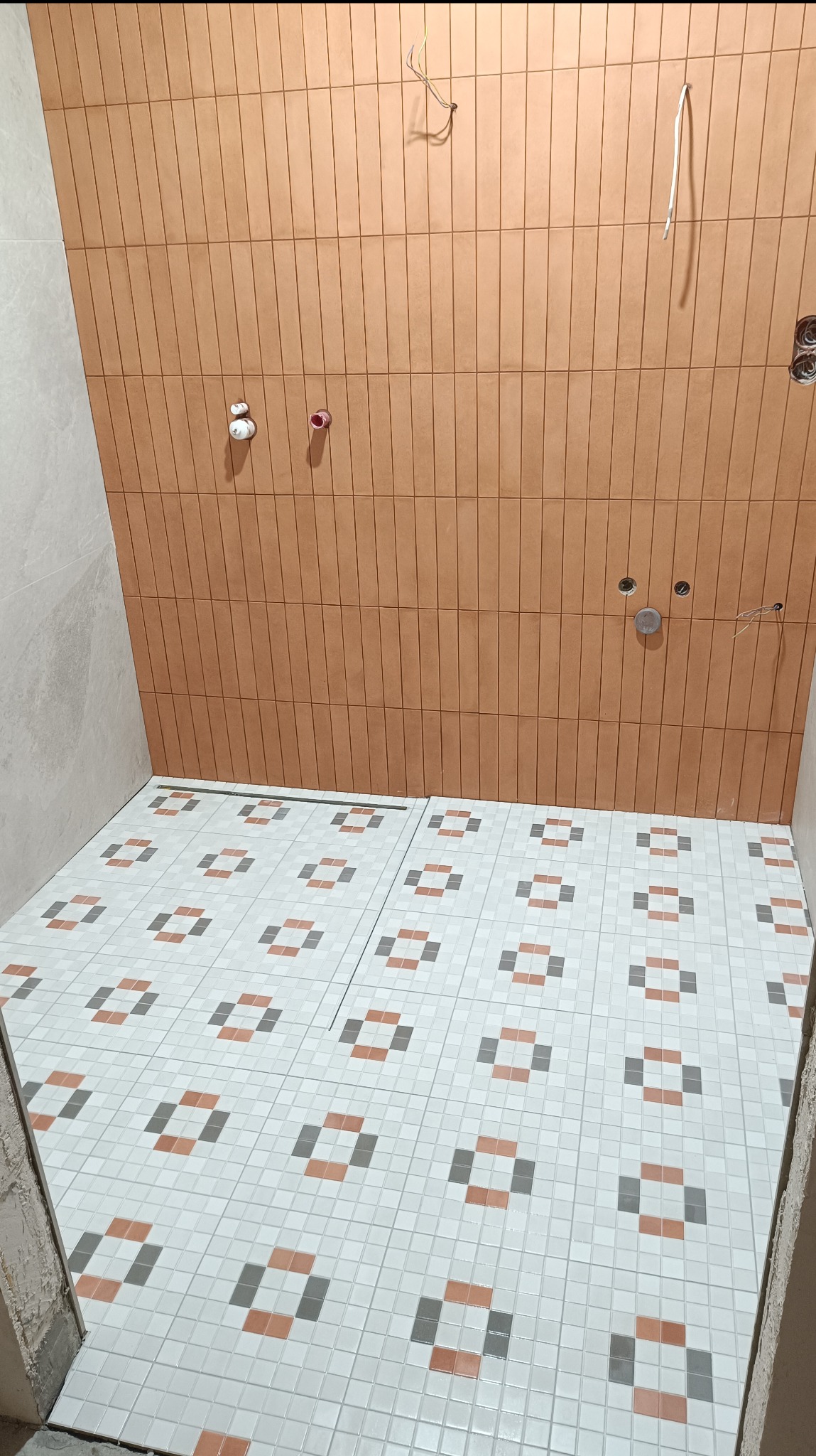 Ściana wyłożona pionowymi, wąskimi, brązowymi płytkami ceramicznymi, z widocznymi otworami na armaturę i wystającymi kablami, oraz podłoga z białych, kwadratowych płytek z wzorem geometrycznym...
