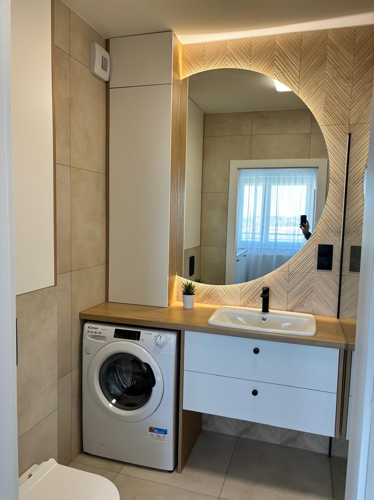 Nowoczesna łazienka z okrągłym lustrem podświetlanym LED, umywalką nablatową z czarną armaturą, białą szafką pod umywalką z dwoma szufladami, pralką Candy i beżowymi płytkami na ścianach...