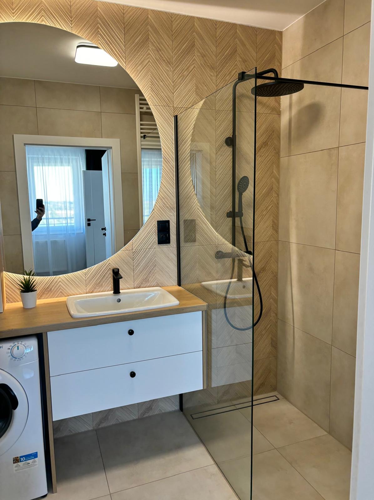 Nowoczesna łazienka z kabiną prysznicową typu walk-in, okrągłym lustrem podświetlanym LED, umywalką nablatową z czarną armaturą i szafką z białymi frontami na tle ściany wykończonej płytkami...