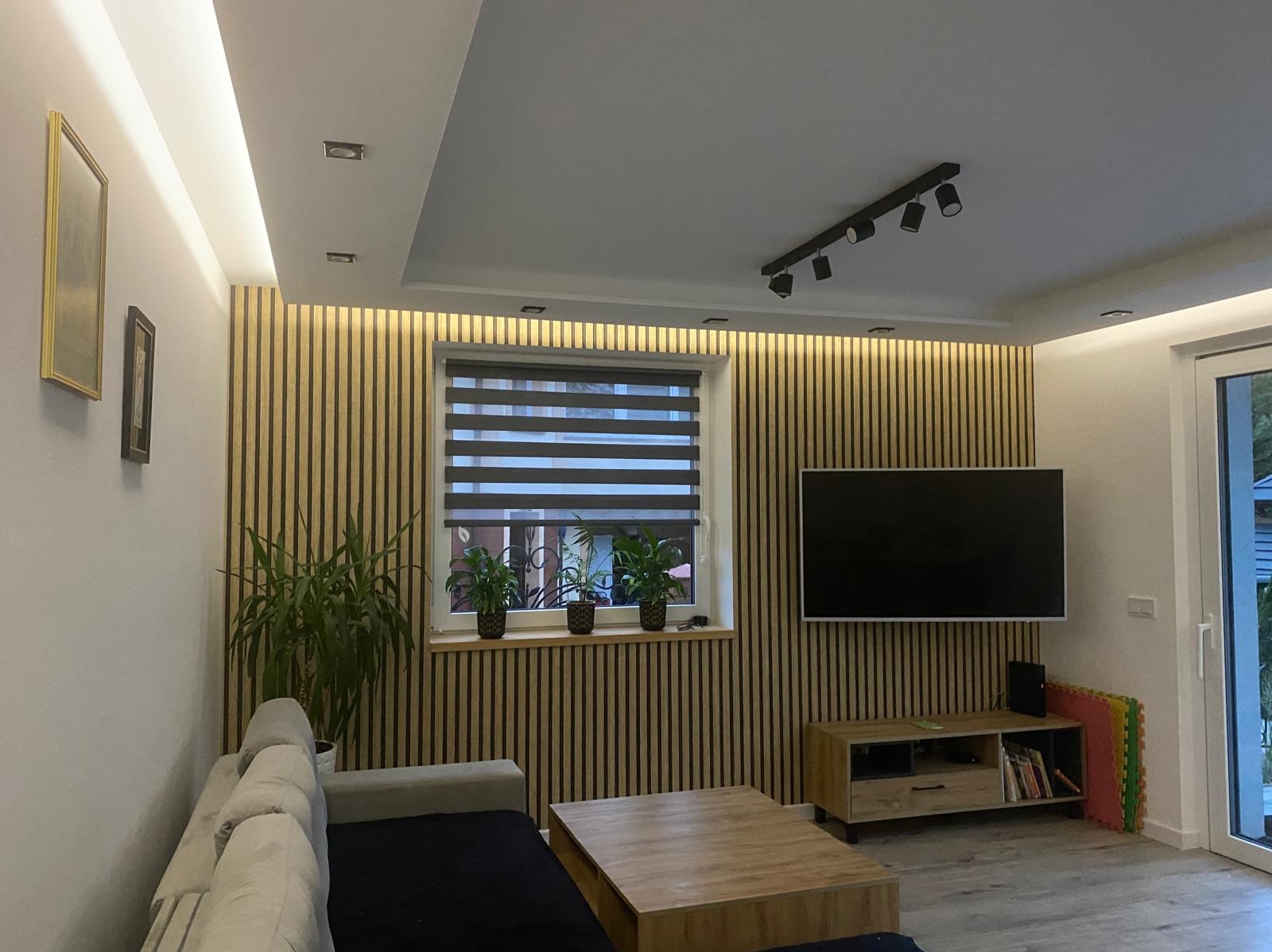 Salon z dekoracyjną ścianą z lameli drewnianych, podświetlonych od góry, z telewizorem na ścianie i roślinami na parapecie, widok z niskiego kąta.