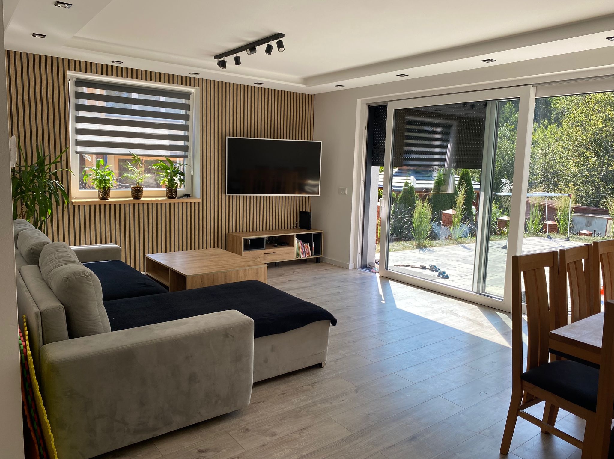 Nowoczesny salon z drewnianą ścianą panelową, telewizorem, szarym narożnikiem i dużym oknem przesuwnym z widokiem na zieleń.