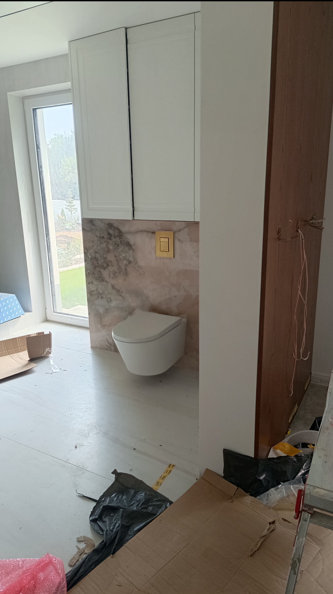 Wisząca toaleta z białym siedziskiem na tle marmurowej ściany z złotym przyciskiem spłukiwania, białe szafki wiszące, widok przez okno na ogród, podłoga częściowo zakryta kartonami i folią.