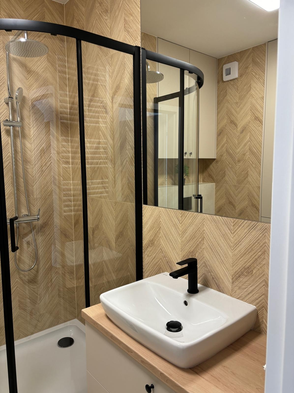 Wykończona łazienka z kabiną prysznicową w czarnej ramie, umywalką nablatową z czarną armaturą i płytkami imitującymi drewno ułożonymi w jodełkę.