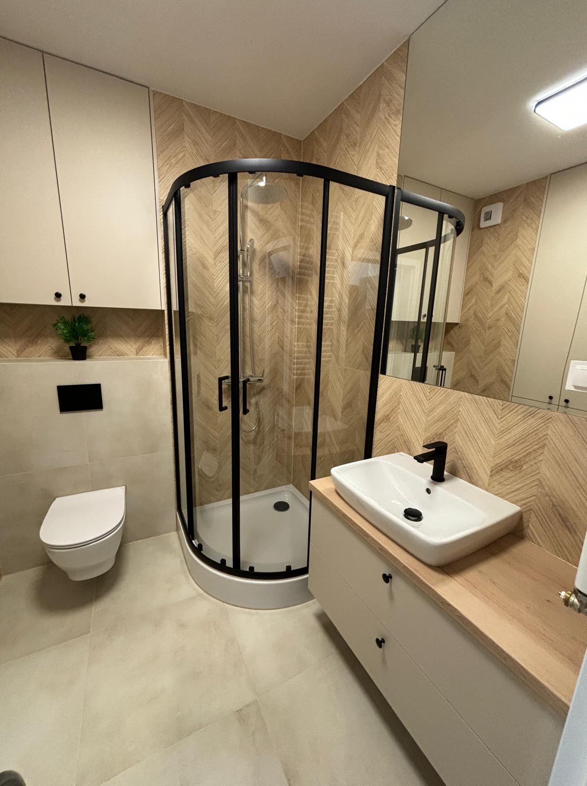 Nowoczesna łazienka z kabiną prysznicową z czarnymi ramami, umywalką nablatową na szafce z drewnianym blatem i toaletą w zabudowie, wykończona płytkami w jodełkę.