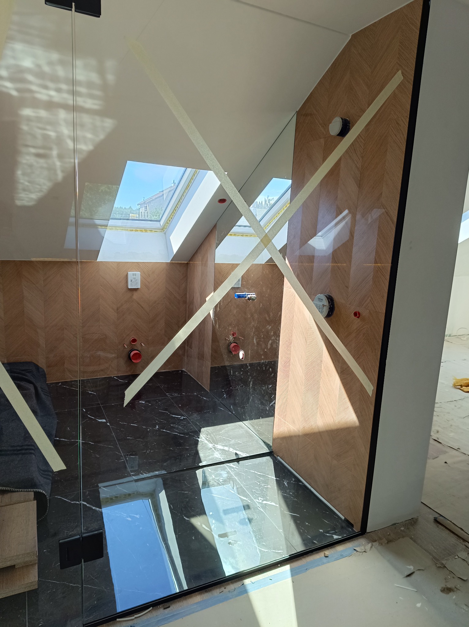Łazienka w trakcie wykańczania: czarne marmurowe płytki podłogowe, ściany wyłożone drewnopodobnymi płytkami w jodełkę, szklana kabina prysznicowa zabezpieczona taśmą malarską, widoczne przyłącza...
