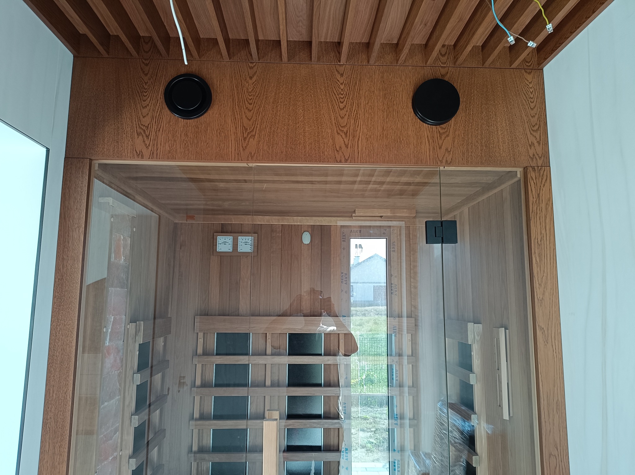 Wnętrze sauny z drewnianą boazerią, przeszklonymi drzwiami i widocznymi elementami wentylacji na suficie.