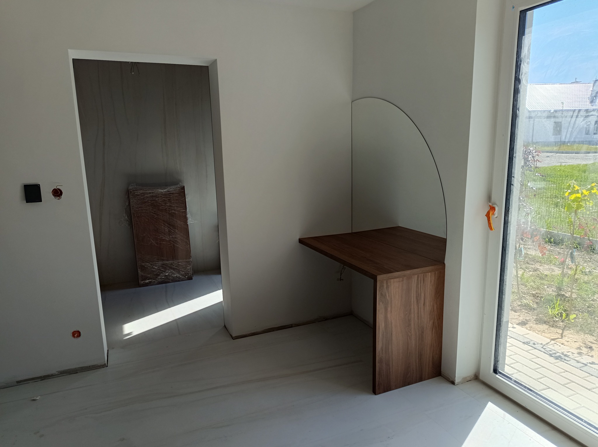 Nowoczesna, drewniana konsola przymocowana do ściany w surowym wnętrzu, białe ściany, duże okno z widokiem na ogród, podłoga z jasnych płytek, drzwi w folii ochronnej w tle.