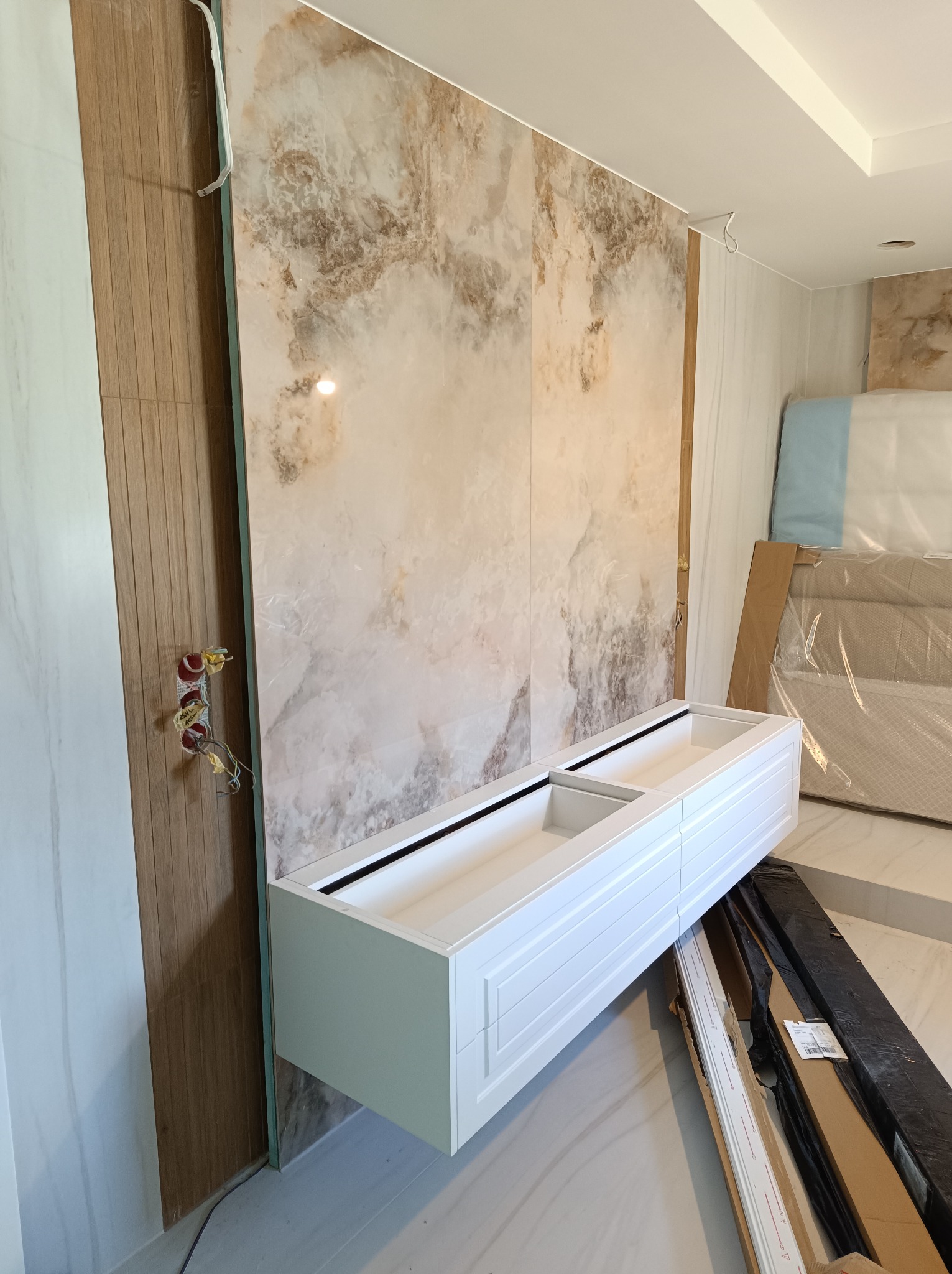 Montaż wiszącej szafki łazienkowej z białymi frontami, na tle marmurowej ściany, widoczne wystające kable elektryczne i elementy wykończeniowe w kartonach.