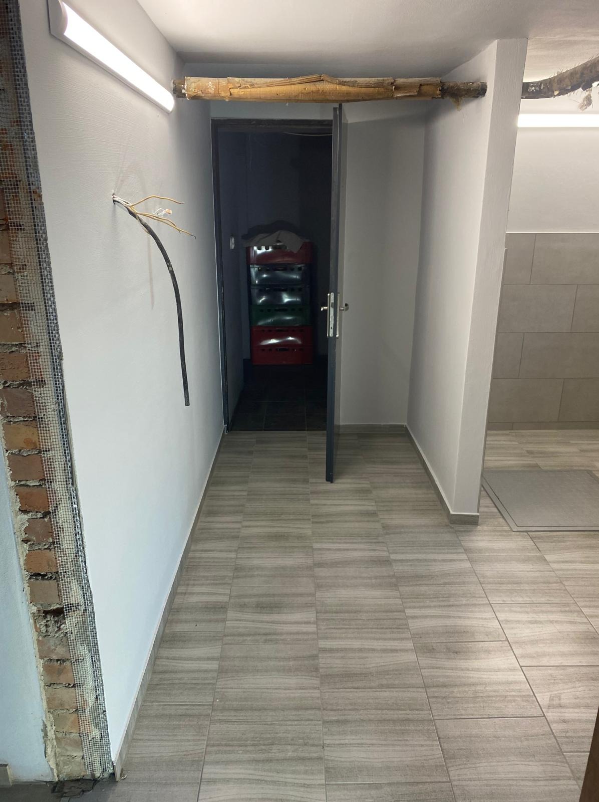 Korytarz w trakcie prac wykończeniowych: ściana z siatką i odsłoniętą cegłą, świeżo pomalowana ściana z wystającymi kablami, płytki podłogowe imitujące drewno, otwarte drzwi do ciemnego...