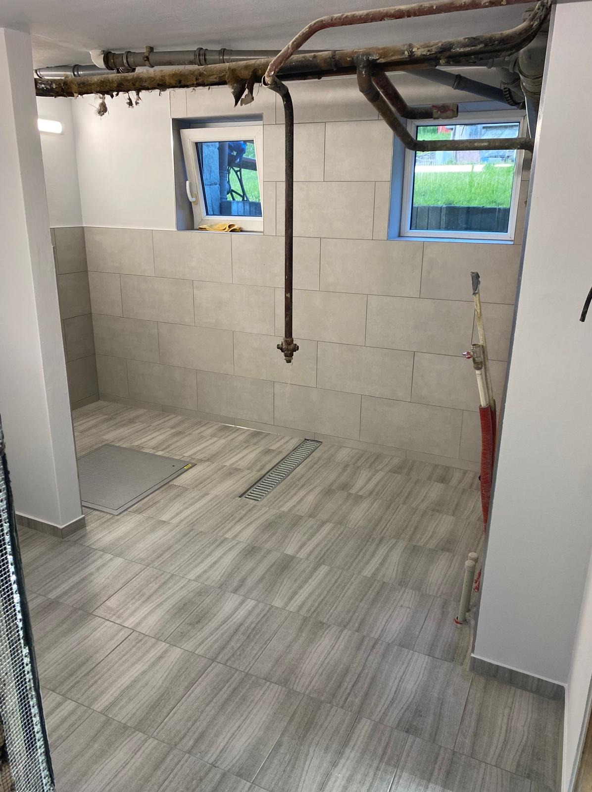 Wykończona łazienka z szarymi płytkami imitującymi drewno na podłodze i ścianach, widoczny odpływ liniowy, rury grzewcze pod sufitem i dwa okna z widokiem na zieleń.