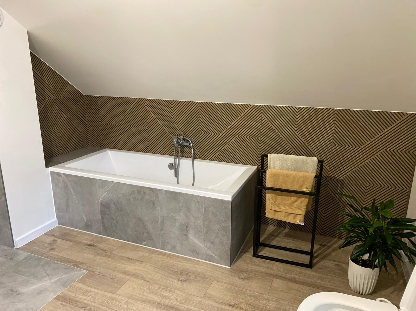 Wykończona łazienka na poddaszu w Kamionce Wielkiej z wanną obłożoną szarymi płytkami imitującymi marmur, dekoracyjną ścianą z geometrycznym wzorem, drewnopodobną podłogą i czarnym stojakiem...