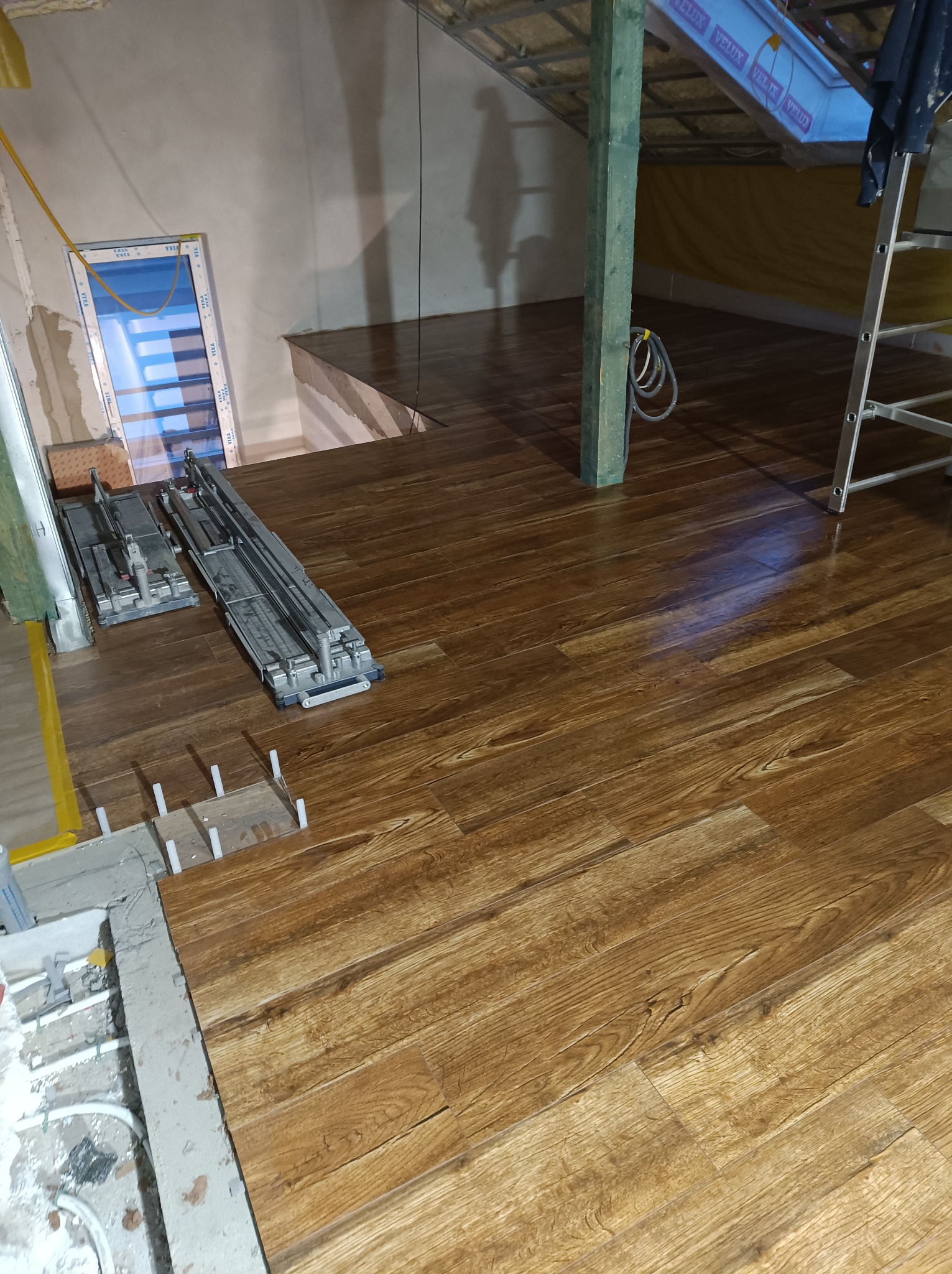 Układanie paneli podłogowych w pomieszczeniu z widocznym oknem dachowym, narzędziami do cięcia i poziomowania, oraz fragmentem instalacji podłogowej.