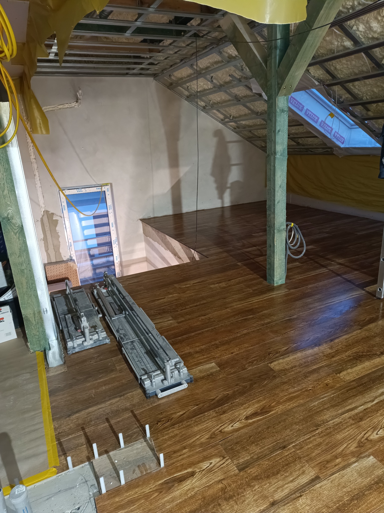 Układanie paneli podłogowych w poddaszu, widoczne narzędzia glazurnicze, białe kliny dystansowe, okno dachowe Velux i drewniany słup konstrukcyjny.