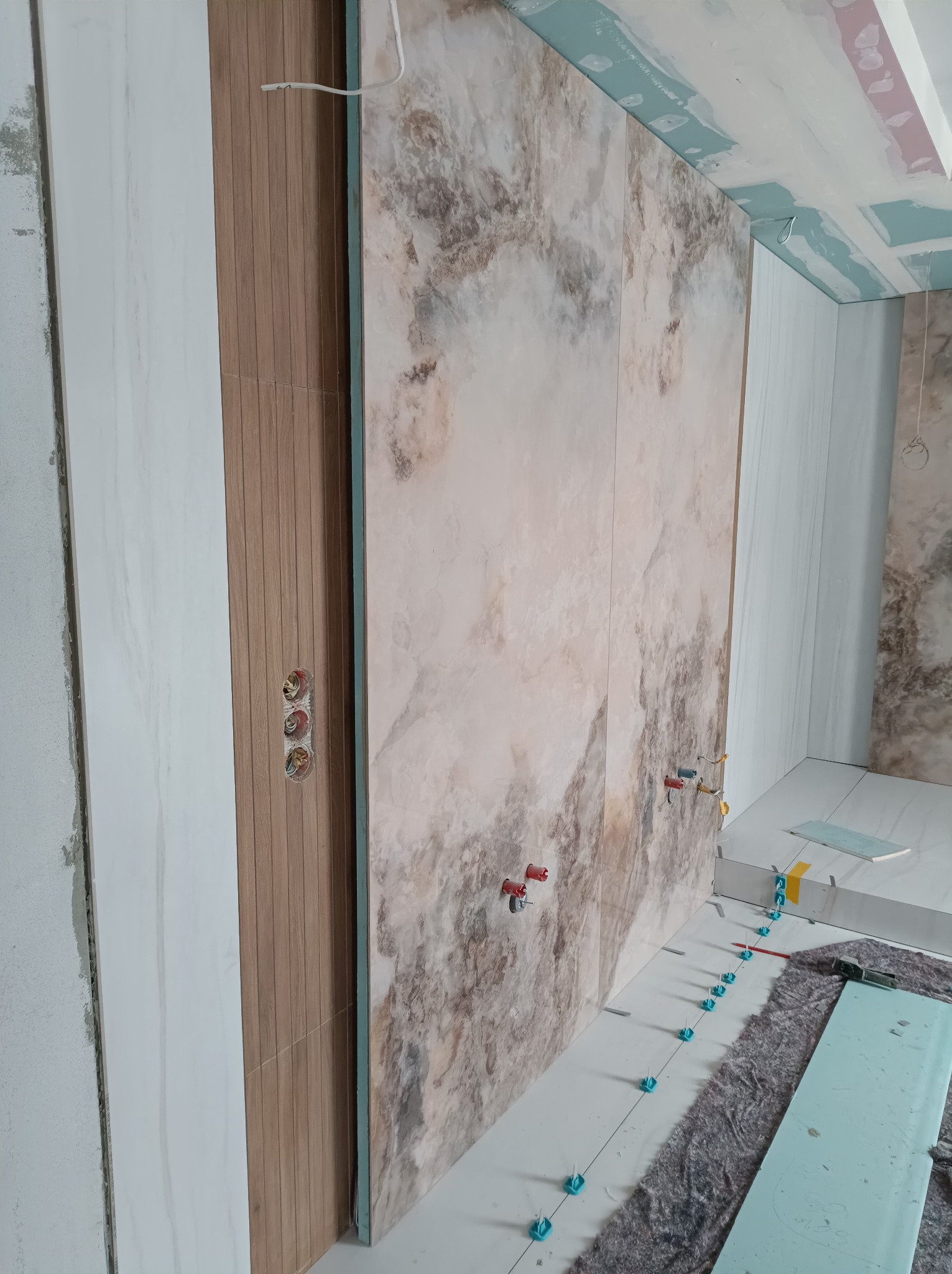 Montaż wielkoformatowych płytek imitujących marmur na ścianie w łazience, widoczne poziomowanie z użyciem klipsów i klinów, surowy stan wykończenia sufitu.