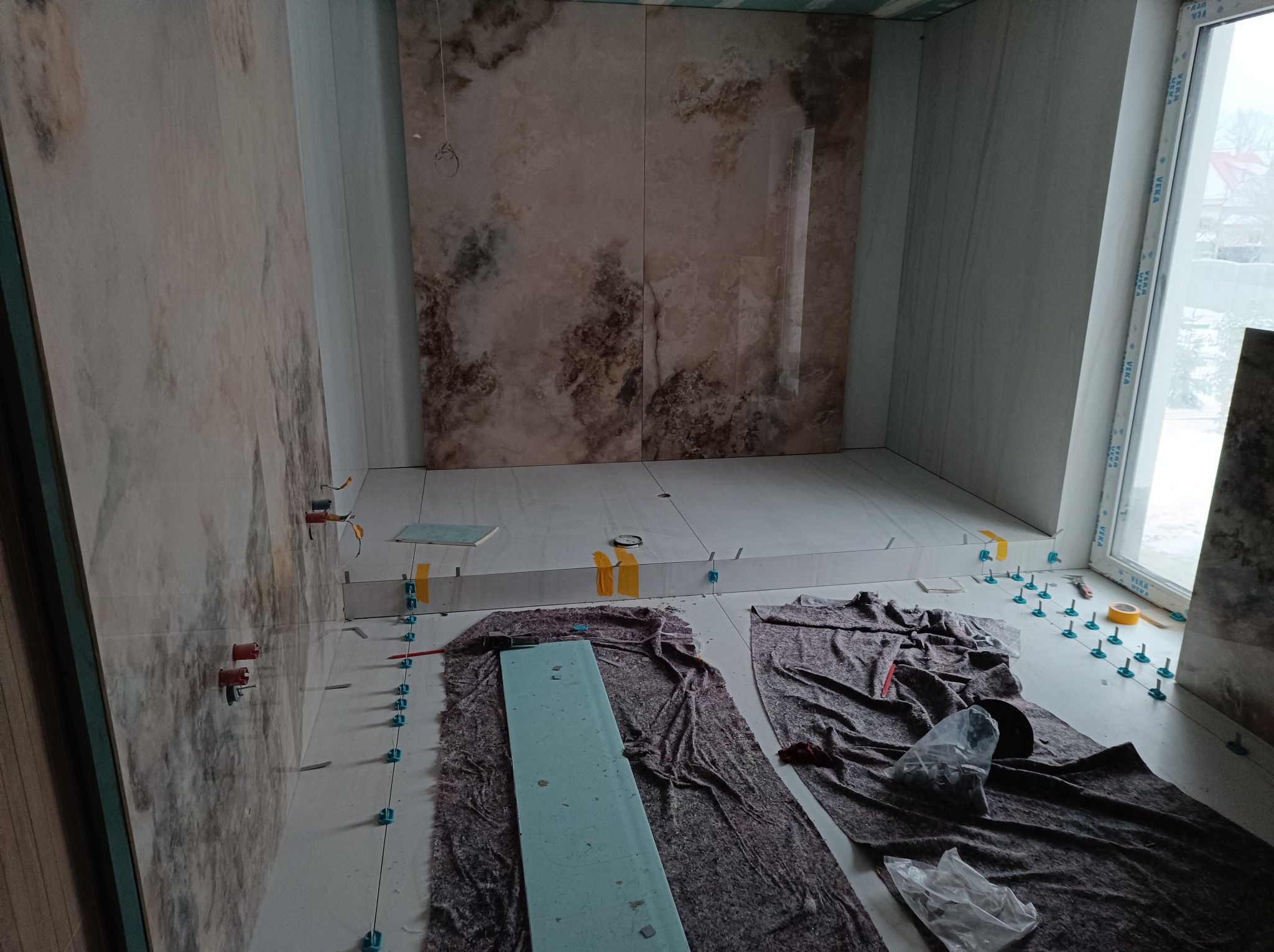 Montaż płytek imitujących marmur w łazience, widoczne narzędzia glazurnicze i poziomica na zabezpieczonej podłodze, ściany częściowo wykończone.