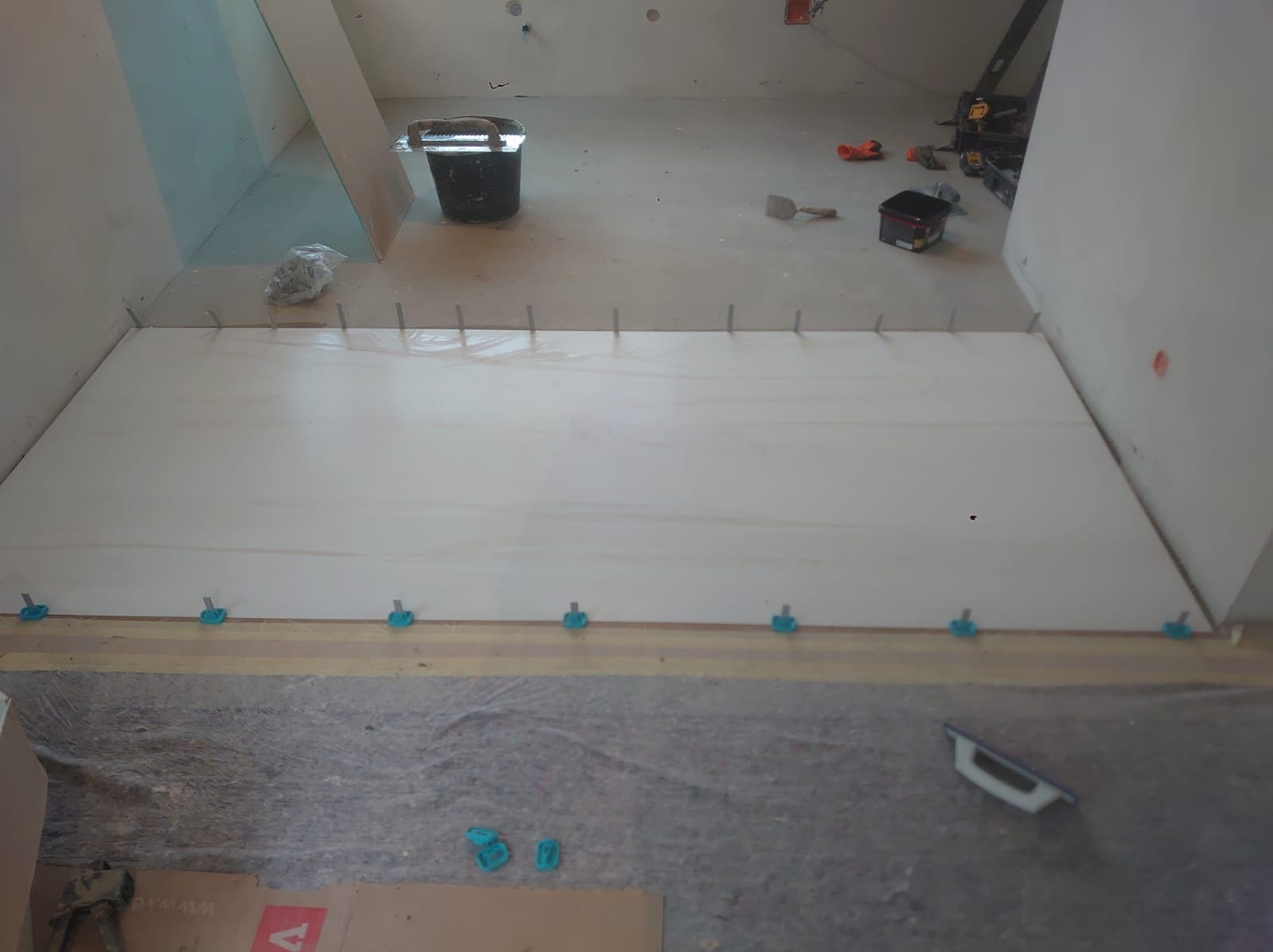 Przygotowana powierzchnia pod płytki podłogowe z użyciem listew wyrównujących i krzyżyków dystansowych, widoczny kubeł z narzędziami i poziomica w tle, surowy stan pomieszczenia.