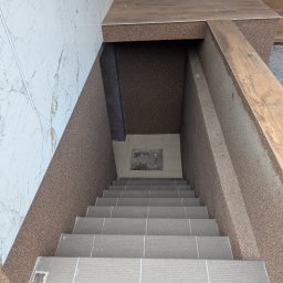 F.H.U. DS Elektryka & Glazura Dariusz Słembarski - Wykończone schody wejściowe z szarych płytek, prowadzące w dół, otoczone ścianami z marmuru i brązowego tynku mozaikowego. Widok z góry.