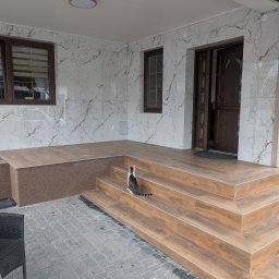 F.H.U. DS Elektryka & Glazura Dariusz Słembarski - Wykończony taras z brązowymi płytkami imitującymi drewno, marmurowe płytki na ścianach i kot siedzący na schodach, dodający uroku.