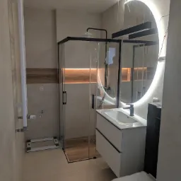 Nowoczesna łazienka po remoncie: kabina prysznicowa z czarnymi ramami, umywalka z szafką, okrągłe lustro z podświetleniem LED i grzejnik.