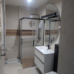 F.H.U. DS Elektryka & Glazura Dariusz Słembarski - Nowoczesna łazienka z kabiną prysznicową typu walk-in, umywalką na szafce i okrągłym lustrem. Beżowe płytki z drewnianym akcentem. Widoczne elementy instalacji.