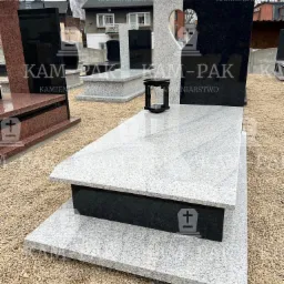 Jasnoszary, warstwowy pomnik z czarnym akcentem i lampionem, na żwirowym podłożu, w tle zabudowa mieszkalna, jeden z pionowych elementów z wyciętym kształtem serca.