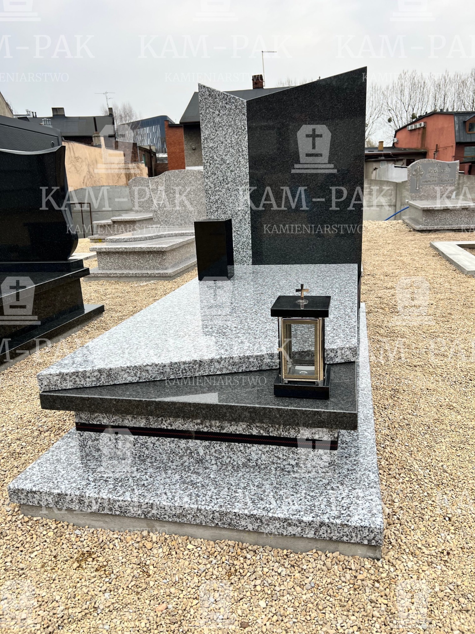 Nowoczesny, dwukolorowy nagrobek z ciemnego i jasnego granitu, z lampionem na płycie, ustawiony na żwirowym podłożu.