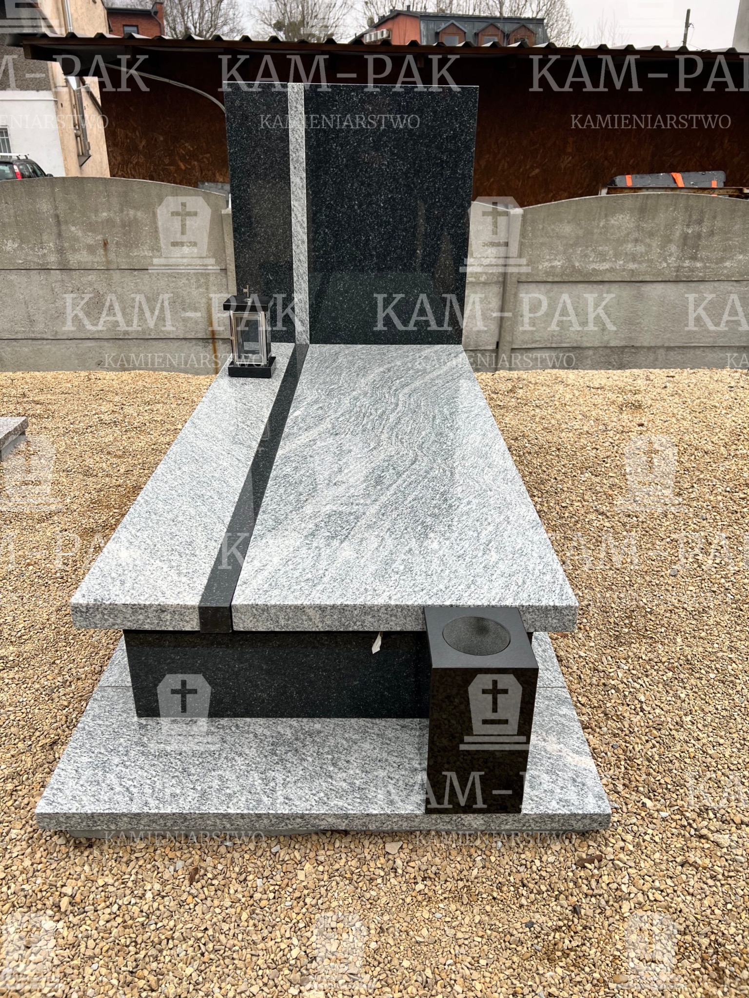 Granitowy pomnik z czarnymi i szarymi elementami, z lampionem i wygrawerowanym krzyżem, ustawiony na żwirowym podłożu.