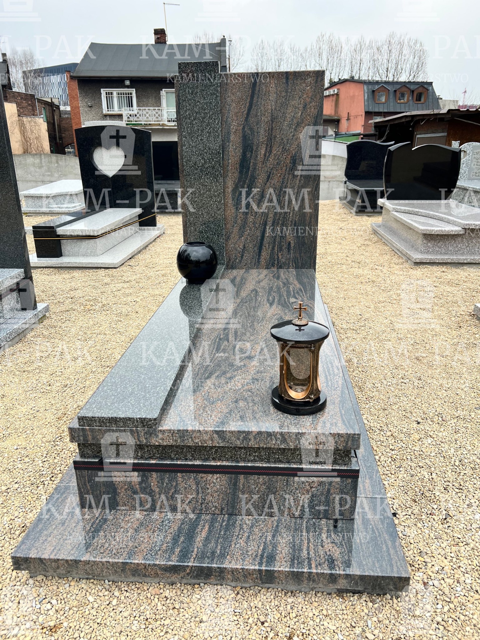 Wypolerowany, dwukolorowy pomnik z granitu z lampionem i wazonem, na żwirowym podłożu, z budynkami w tle.