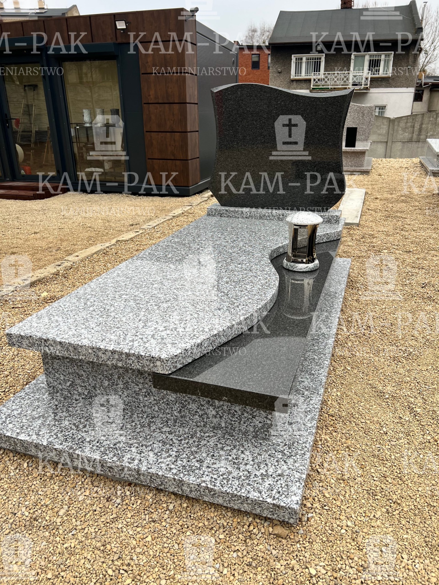 Granitowy nagrobek z ciemnoszarą tablicą z symbolem krzyża i lampionem, na żwirowym podłożu, z budynkami w tle.