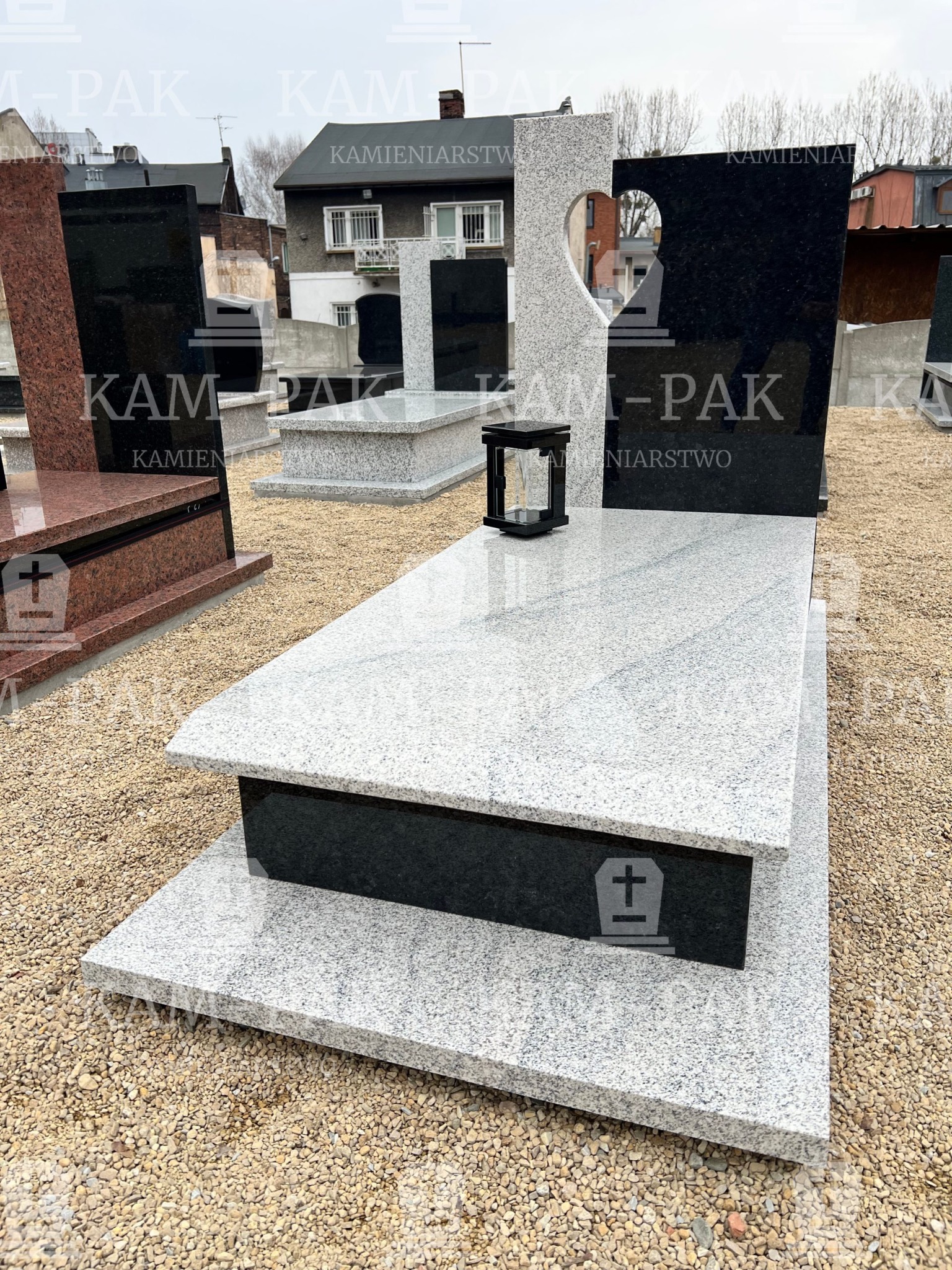 Jasnoszary, warstwowy pomnik z czarnym akcentem i lampionem, na żwirowym podłożu, w tle zabudowa mieszkalna, jeden z pionowych elementów z wyciętym kształtem serca.