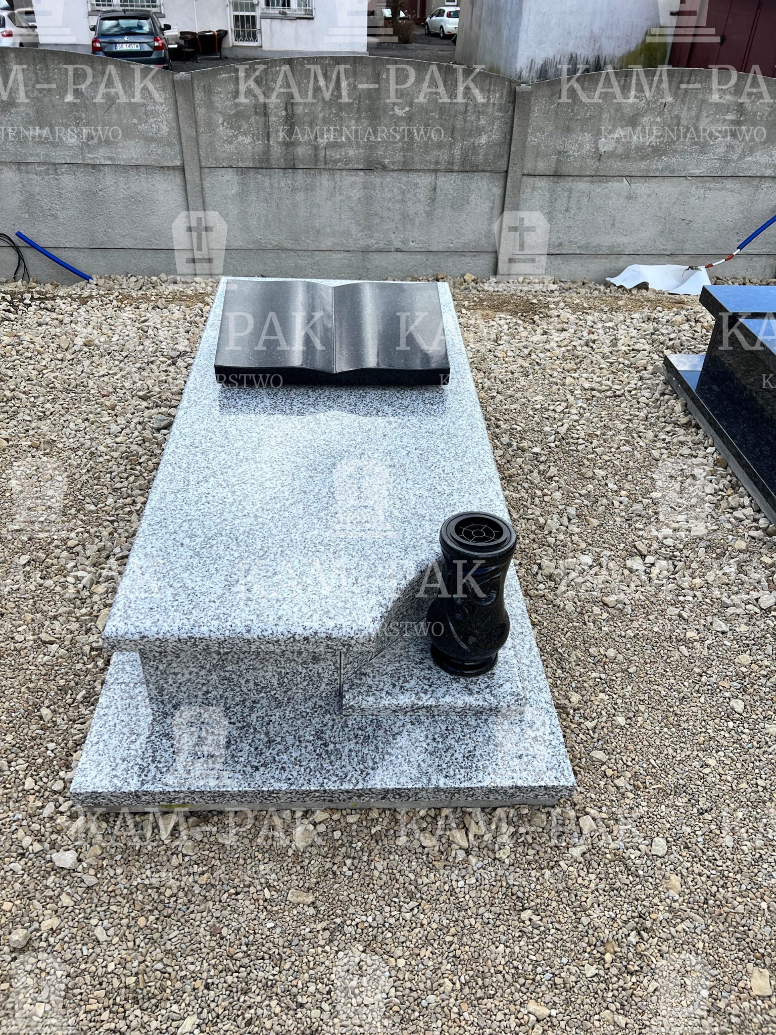Jasnoszary, granitowy pomnik z czarną, otwartą księgą i wazonem na żwirze, na tle betonowego ogrodzenia z widocznym logo firmy kamieniarskiej.