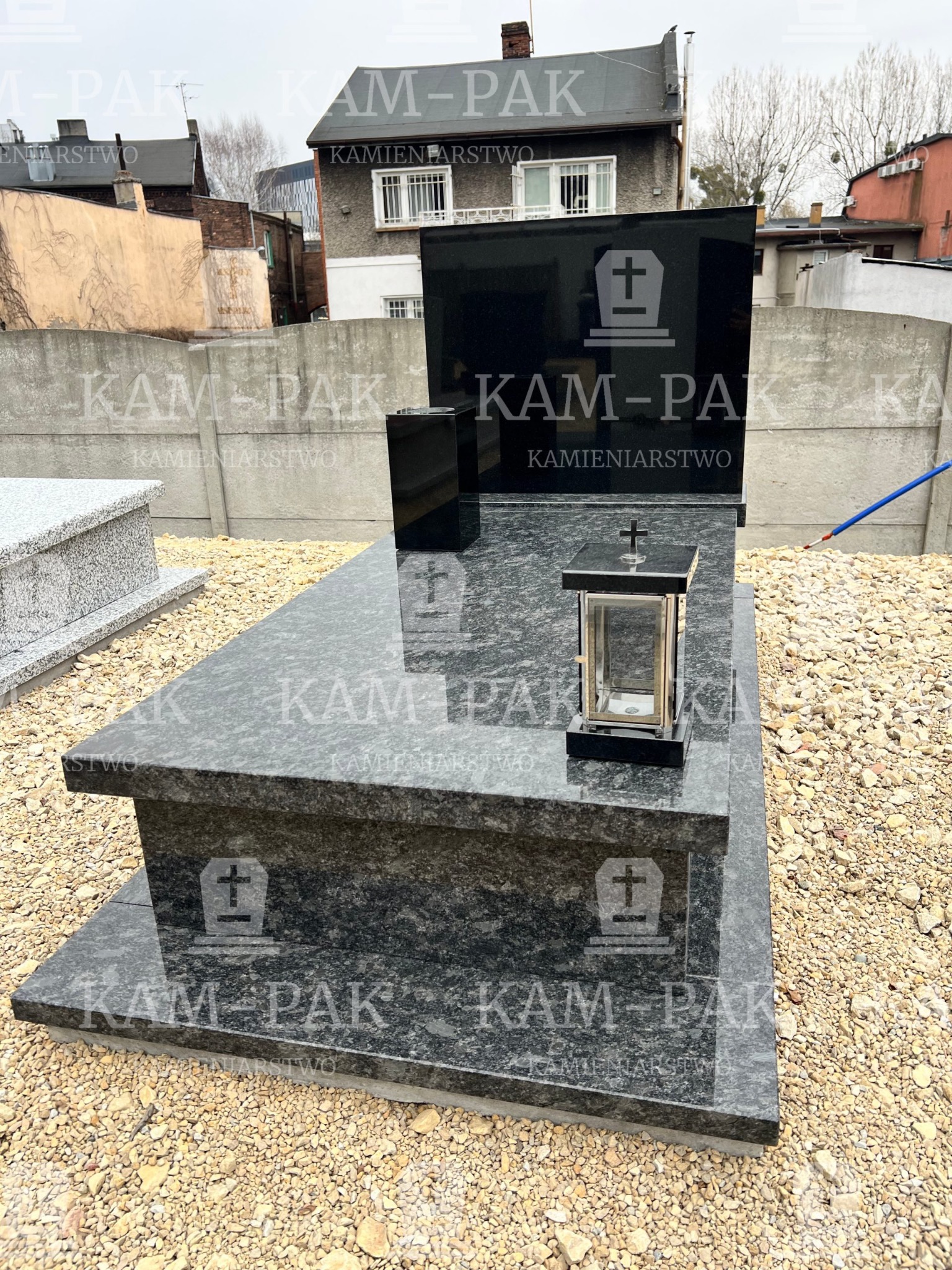 Czarny, polerowany pomnik z szarego granitu z lampionem i wazonem, na tle budynków mieszkalnych i betonowego ogrodzenia.