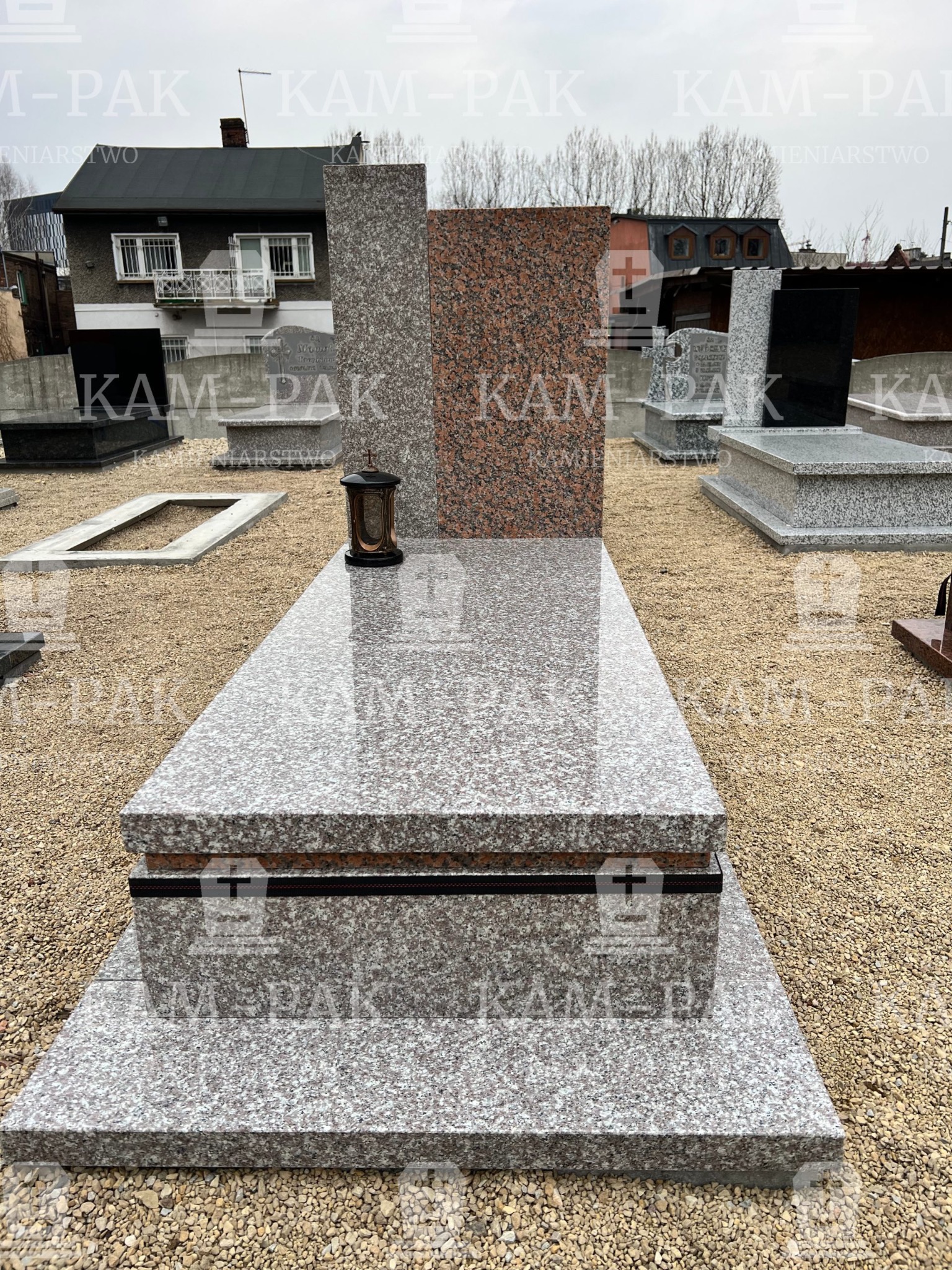 Jasnoszary granitowy pomnik z brązowym lampionem, na tle innych pomników i zabudowań.