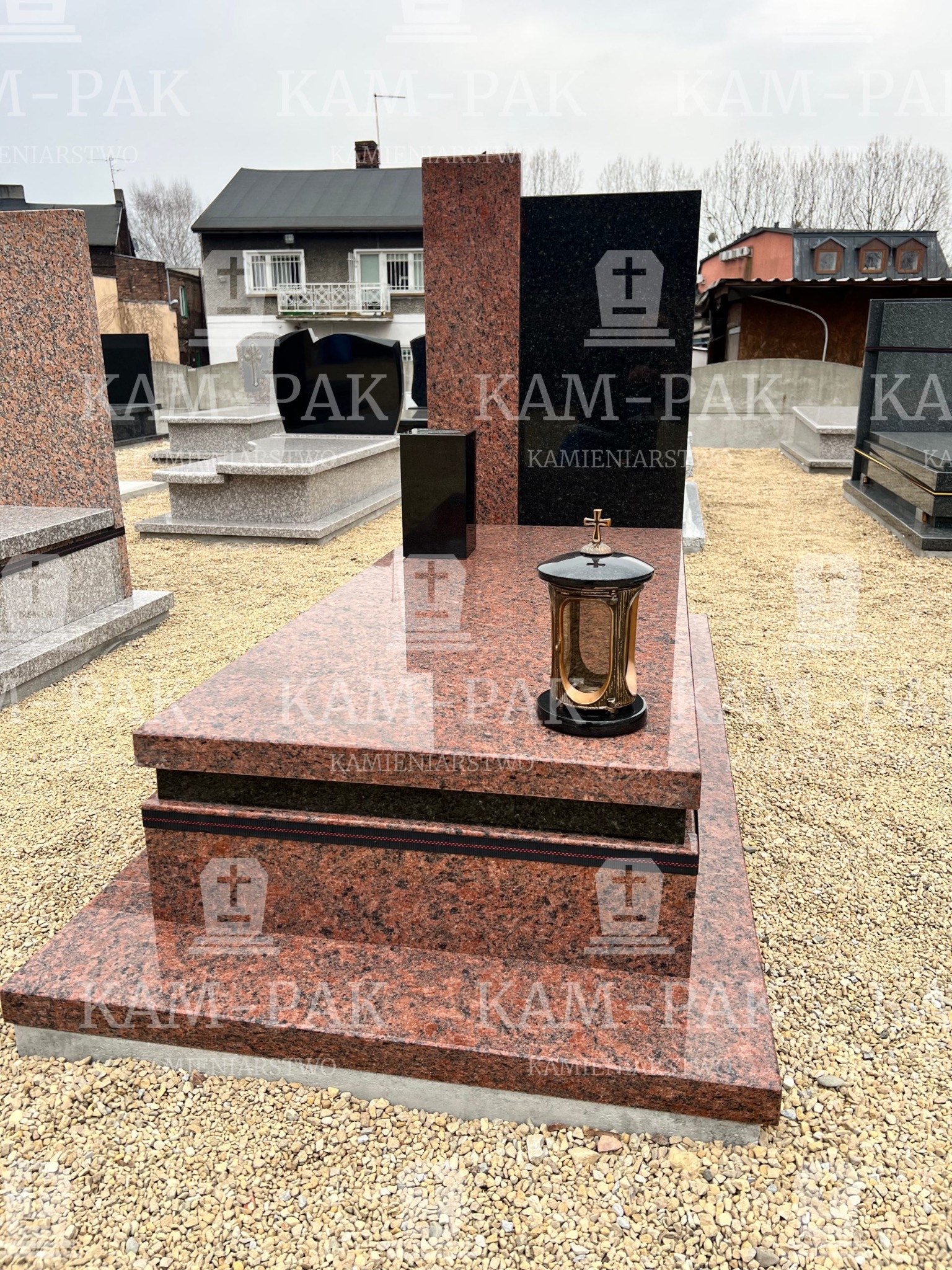 Czerwono-czarny, dwuelementowy pomnik z polerowanego granitu z lampionem i wazonem, ustawiony na żwirowym podłożu, na tle innych nagrobków i budynku.