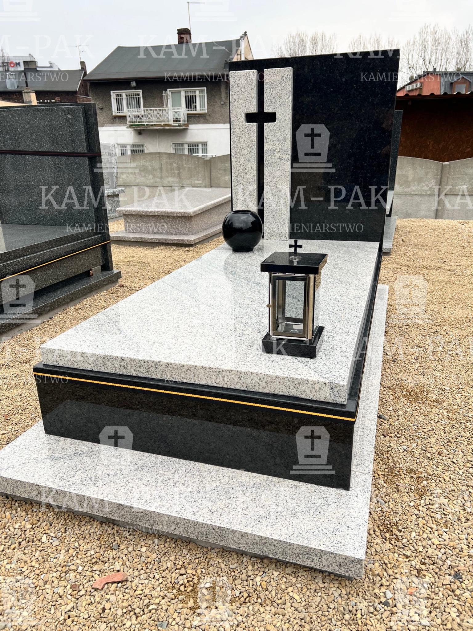 Czarno-biały pomnik z krzyżem i lampionem na cmentarzu, widok z boku, tło stanowią budynki mieszkalne.